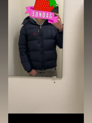 Mörkblå pufferjacka från Polo Ralph Lauren - Snygg mörkblå pufferjacka från Polo Ralph Lauren med huva och dragkedja framtill. Jackan har det klassiska röda Polo-logot på bröstet och en vadderad design som håller dig varm under kalla dagar. Perfekt för dig som vill ha både stil och funktion.