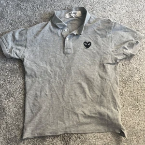 Grå pikétröja från Comme des Garçons Play - Grå pikétröja med klassisk krage och knäppning framtill. Svart hjärta-logga broderad på bröstet. Perfekt för en stilren och avslappnad look. Storleken är L men sitter mer som en M, tveka inte på att skriva om frågor, priset är låg eftersom den passa inte mig så bra och vill sälja snabbt
