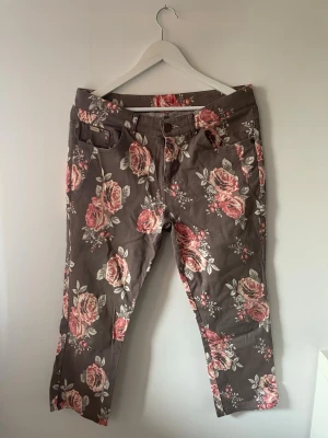 Gråblommiga jeans med rosor - Brun/grå jeans med rosa och vita rosor i all-over print. Modellen har klassisk femficksdesign, dragkedja och knapp i midjan. Jeansen är i ett mjukt bomullstyg och har croppad längd. 