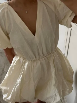 Kort ljus klänning med v-ringning - Supersöt kort klänning i ljus beige färg med djup v-ringning och puffiga korta ärmar. Klänningen har markerad midja med resår och voluminös kjol. Perfekt för varma dagar och enkel att styla med smycken.