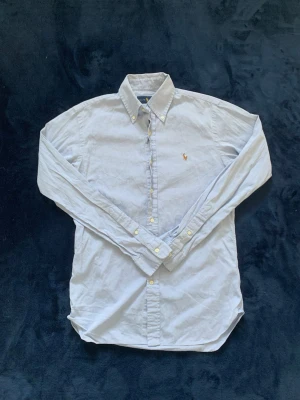 Ljusblå skjorta från Ralph Lauren - Klassisk ljusblå skjorta från Ralph Lauren med broderad logga på bröstet. Skjortan har knappar hela vägen, button-down krage och långa ärmar. Perfekt för en clean och stilren look. 10/10 skick.