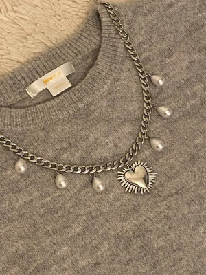 Silverfärgat halsband med pärlor & hjärta - Stilrent halsband i silverfärgad metall med droppformade pärlor och ett stort hjärthänge med solstråle-design. Kedjan är chunky och ger en cool vibe, perfekt för att lyfta din outfit med en unik detalj.
