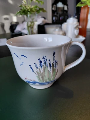 Vit kopp med blått blommotiv - Säljer en vit porslinskopp med handtag, dekorerad med blå lavendel-liknande blommor och gröna blad samt några blå fåglar. Koppen har en klassisk form med lätt utsvängd kant och rymmer gott om dryck. Perfekt för te eller kaffe.