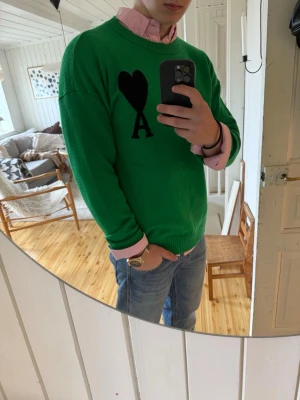 Grön stickad tröja från Ami Paris - Snygg grön stickad tröja från Ami Paris med svart hjärta och bokstaven A på bröstet. Tröjan har rund halsringning, ribbade muddar och en avslappnad passform. Perfekt att bära över en skjorta för en trendig look.