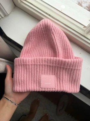  Acne mössa - Rosa ribbstickad mössa från Acne Studios med uppvikt kant och det ikoniska ansikts-emblemet framtill. Mjuk och varm, perfekt för kalla dagar. Materialet är ullblandning som ger både komfort och stil.