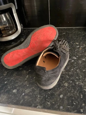 Svarta Christian Louboutin sneakers - Säljer ett par svarta sneakers från Christian Louboutin med klassisk röd sula och coola nitar framtill. Skorna är i mocka och har svarta skosnören. Insidan är i ljust läder för extra komfort. Perfekta för dig som vill sticka ut med din stil. Till bra pris 