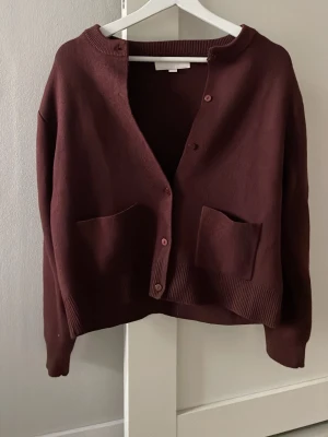 Vinröd kofta med fickor - Mysig vinröd kofta med rund halsringning och knäppning framtill. Två stora fickor framtill och ribbade muddar vid ärmslut och nederkant. Oanvänd och ny.
