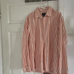 Randig skjorta från H&M i orange och vit - Säljer en snygg randig skjorta från H&M i orange och vitt. Skjortan har klassisk krage, långa ärmar och knäppning framtill. Regular fit och tillverkad i bomull, perfekt för en chill och stilren look.