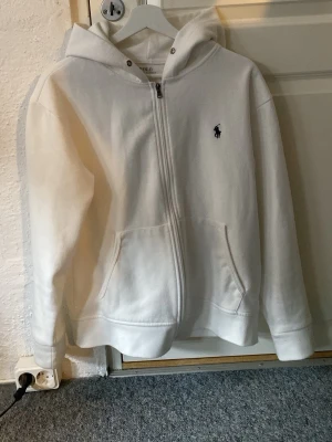Zip Polo Ralph Lauren - Säljer en stilren vit hoodie från Polo Ralph Lauren med dragkedja framtill och två fickor. Klassisk huva, saknar  dock snörning, och den ikoniska lilla loggan broderad på bröstet. Perfekt för en clean och avslappnad look. Använd en gång, nyskick