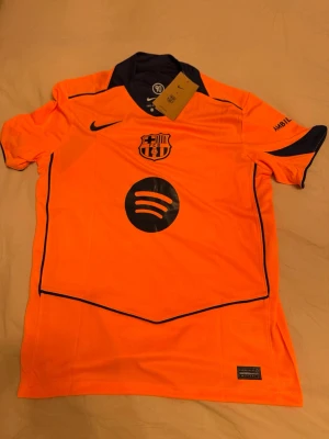 FC Barcelona orange matchtröja Nike - Snygg orange FC Barcelona matchtröja från Nike med mörkblå detaljer och klubbmärke på bröstet. Spotify-logga framtill och UNHCR-tryck bak. Nummer 10 och Lamine Yamal på ryggen. Perfekt för dig som älskar fotboll och vill sticka ut på planen.Priset kan diskuteras vid snabba affärer.