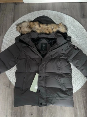 Svart dunjacka från Canada Goose - Snygg svart dunjacka från Canada Goose med stor huva och pälskant i naturfärg. Jackan har flera fickor framtill, patch med logga på ärmen och kraftig dragkedja. Perfekt för kalla vinterdagar och riktigt varm.
