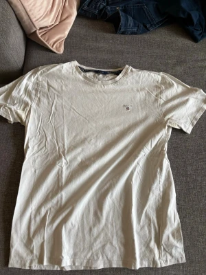 Vit t-shirt från Gant med logga - Säljer en klassisk vit t-shirt från Gant med liten broderad logga på bröstet. T-shirten har rund halsringning och korta ärmar. Perfekt för dig som gillar stilrena basics och vill ha något som funkar till jeans eller shorts.