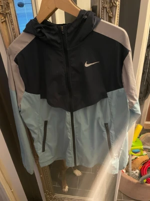 Nike miller (nice för sommaren + hösten) - Nike vindjacka i färgblockad design med mörkblå, ljusblå och grå toner. Jackan har huva, hel dragkedja framtill och två fickor med dragkedja. Tillverkad i lätt och vindtätt material, perfekt för blåsiga dagar. Ikonisk Nike-logga på bröstet.