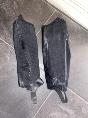 Svarta shortchaps i läder och tyg - Ett par svarta shortchaps med dragkedja och resår. De är tillverkade i en kombination av slitstarkt tyg och glansigt läder, vilket ger både stil och funktion. Perfekta som komplement till ridstövlar och har en sportig look med praktiska detaljer. Tvättas och är som nya innan du får dem.