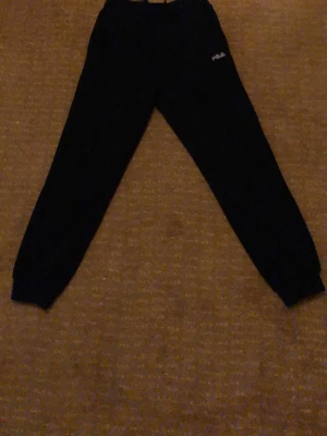 Svarta mjukisbyxor från FILA - Säljer ett par svarta mjukisbyxor från FILA med logga på ena benet. Byxorna har resår i midjan och vid bensluten för en skön och avslappnad passform. Perfekta för chill eller träning. Materialet känns mjukt och bekvämt.