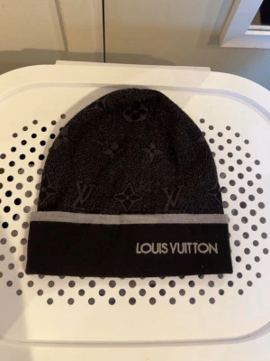 Monogram mössa - Snygg svart mössa  med grå kant och broderad logga framtill. Mössan har diskret monogrammönster över hela ytan och är tillverkad i ett mjukt, stickat material. Perfekt accessoar för att lyfta din stil.