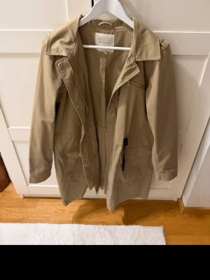 Beige parkas från Selected Femme - Stilren beige parkas från Selected Femme med dragkedja och tryckknappar framtill. Jackan har justerbar midja med snörning, stora fickor och en klassisk krage. Tillverkad i ett slitstarkt bomullsmaterial som passar perfekt för kyligare dagar.