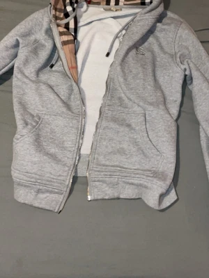 Grå zip-hoodie från Burberry utan dragkedja  - Snygg grå hoodie med dragkedja från Burberry. Hoodien har klassiskt rutigt foder i huvan, dragsko och två fickor framtill. Tillverkad i mjukt bomullsmaterial och har ribbade muddar vid ärmslut och nederkant. Perfekt för en stilren och avslappnad look.