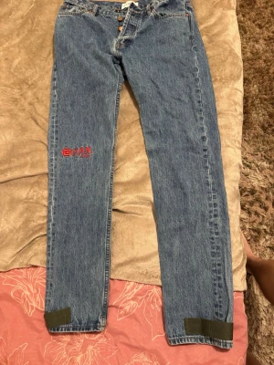 Blå jeans från Han Kjøbenhavn - Säljer ett par blå raka jeans från Han Kjøbenhavn med röd brodyr på vänster ben och gröna detaljer vid bensluten. Klassisk femficksmodell med knappgylf och robust denimkänsla. Perfekta för dig som gillar streetwear och unika detaljer.