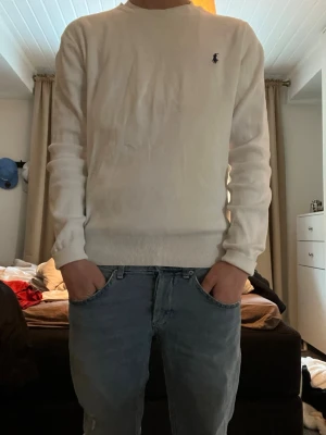 Vit stickad tröja från Ralph Lauren - Klassisk vit stickad tröja från Ralph Lauren med rund halsringning och diskret broderad logga på bröstet. Tröjan har långa ärmar och ribbade muddar vid ärmslut och nederkant. Perfekt till jeans för en clean och stilren look.