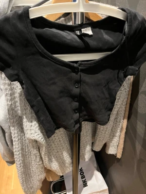 Svart u-ringad crop top med knappar - Snygg svart crop top från Bik Bok med knappar framtill. Toppen har en rund halsringning och är gjord i ett mjukt, stretchigt material som sitter tajt. 