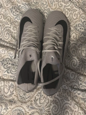 Gråa Nike Mercurial Elite fotbollsskor - Snygga gråa Nike Mercurial fotbollsskor med svart swoosh på ovansidan. Skorna har snörning och en strömlinjeformad design för bra passform och kontroll på planen. Tillverkade i ett lätt syntetmaterial som ger bra känsla och grepp på konstgräs och gräs. 