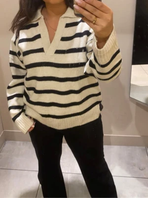 Randig stickad tröja med krage - Snygg stickad tröja i vitt med svarta horisontella ränder och en djup v-ringning med krage. Tröjan har långa ärmar och ribbade muddar vid ärmslut och nederkant. Perfekt för en avslappnad och trendig look. Den är storlek S men passar bättre till M, så lite stor i storleken