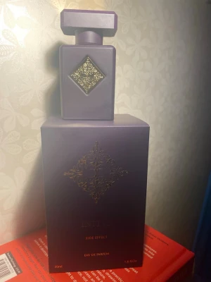 Initio Side Effect Eau de Parfum - Initio Side Effect Eau de Parfum är en lyxig parfym i en elegant mörklila flaska med guldfärgad dekor. Flaskan har en fyrkantig form och ett distinkt geometriskt lock. Förpackningen matchar flaskans färg och har samma guldfärgade mönster. Volymen är 50 ml.