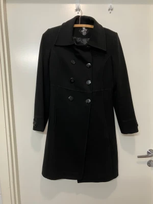 Svart dubbelknäppt kappa Green Cats - Stilren svart kappa från Green Cats i storlek 42. Klassisk dubbelknäppning med stora svarta knappar och bred krage. Lång modell med markerad midja och knappdetaljer vid ärmslut. Perfekt för kyligare dagar och enkel att matcha med olika outfits. 