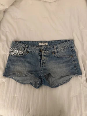 Lågmidjade blå jeansshorts med paljettdetaljer - Säljer ett par blå jeansshorts från Global med paljettdetaljer på ena framfickan och bakfickan. Klassisk femficksmodell med knapp och dragkedja. Perfekta för sommaren och har en avslappnad passform.