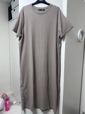 Beige lång t-shirtklänning  - En enkel och stilren beige långklänning med rund halsringning och korta ärmar. Klänningen har en rak passform och är tillverkad i ett mjukt, strukturerat material som känns bekvämt mot huden.
