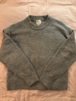 Grå stickad tröja med paljetter - Mysig grå stickad tröja med rund hals och långa ärmar. Tröjan har små silverfärgade paljetter insydda över hela plagget för en extra touch. Ribbstickade muddar vid ärmslut och nederkant. Perfekt för dig som gillar en enkel men ändå glittrig stil.