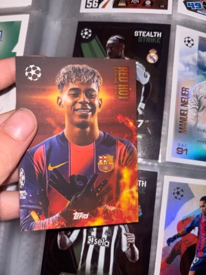 Match Attax Red Hot Lamine Yamal - Ultra-sällsynt Red Hot Lamine Yamal samlarkort från Topps Match Attax Champions League. Kortet har en röd och orange eld-design, FC Barcelona-logga och Champions League-symbol. Perfekt för dig som gillar fotboll och samlarkort.