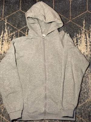 Grå zip hoodie från Jack & Jones - Snygg grå hoodie från Jack & Jones med dragkedja framtill och två fickor. Tillverkad i mjukt sweatshirtmaterial och har en klassisk huva. Perfekt för en avslappnad och chill stil.