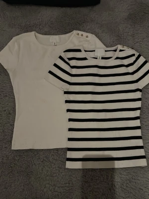 Två kortärmade toppar/ t-shirts  - Två tajta toppar från H&M, en helvit och en vit med svarta ränder. Båda har korta ärmar och dekorativa guldfärgade knappar på axeln. Perfekta för en enkel och snygg look. Materialet känns mjukt och bekvämt (sticks ej). Använda några ggr styck, lite knoppriga men annars inga tecken på användning. Säljer då ja ej använder de:))