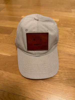 Beige keps med brun patch från Acne Studios - Säljer en beige keps från Acne Studios med den ikoniska bruna face-patchen framtill. Kepsen har klassisk böjd skärm och justerbar rem bak. Tillverkad i slitstark bomullscanvas, perfekt för en clean och stilren look.