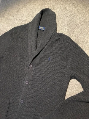 Mörkgrå kofta från Polo Ralph Lauren - Mysig mörkgrå stickad kofta från Polo Ralph Lauren med sjalkrage och knäppning framtill. Klassisk broderad logga på bröstet och två fickor. Perfekt för lager på lager och chill stil.