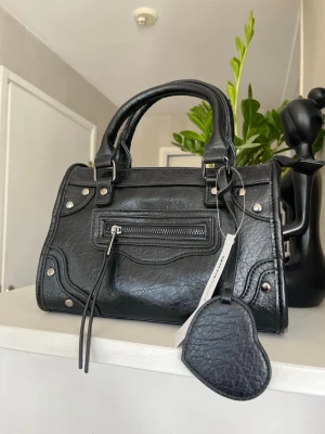 Svart handväska med hjärtdetalj - Stilren svart handväska från Gina Tricot i veganskt läder med silverfärgade nitar och dragkedja framtill. Väskan har dubbla handtag och en söt hjärtformad detalj. Perfekt storlek för det viktigaste och passar till många outfits.