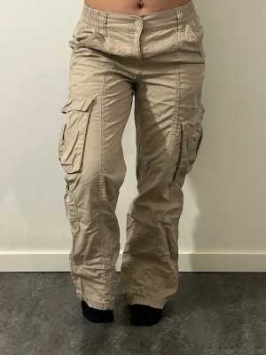 Beiga cargobyxor med fickor - Snygga beiga cargobyxor med flera stora fickor på sidorna och avslappnad passform. Byxorna har resår i midjan och klassisk cargostil. Perfekta för en chill och trendig look. Materialet är bomull och de har knapp- och dragkedjestängning.