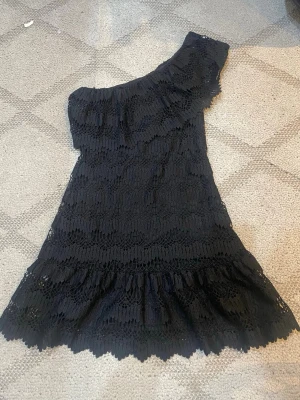 Svart spetsklänning oneshoulder - Snygg svart klänning i spets med oneshoulder-design och volangdetaljer upptill och nertill. Klänningen har en kort längd och ett elegant mönster i spets som ger en trendig look. Perfekt för dig som vill sticka ut med en unik stil.