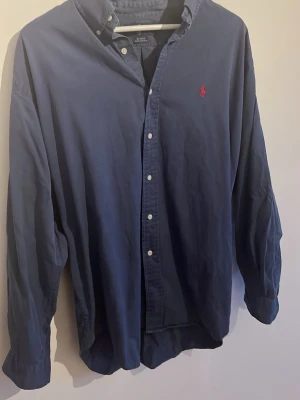 Marinblå skjorta från Ralph Lauren - Klassisk marinblå skjorta från Ralph Lauren med röd broderad logga på bröstet. Skjortan har knappar framtill, button-down krage och långa ärmar. Tillverkad i mjuk bomull som känns skön mot huden. Perfekt för en stilren och avslappnad look. OBS relativt stor i storlek!