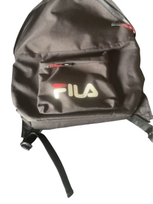 Svart ryggsäck från FILA - Klassisk svart ryggsäck från FILA med stor logga på den främre fickan. Ryggsäcken har en stor huvudficka och en mindre ytterficka med dragkedja i rött. Justerbara axelband och enkel, stilren design i slitstarkt tyg.
