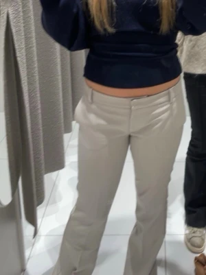 Beiga kostymbyxor med utsvängda ben - Snygga beiga kostymbyxor med utsvängda ben och låg midja. Byxorna har klassisk dragkedja och knapp framtill samt diskreta fickor. Perfekta för en stilren look och lätta att matcha med olika toppar.