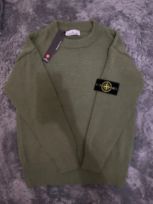 Olivgrön stickad tröja Stone Island - Snygg olivgrön stickad tröja från Stone Island med klassisk rund halsringning och ribbade muddar. Tröjan har den ikoniska Stone Island-loggan på vänster ärm och är tillverkad i mjukt stickat material. Perfekt för dig som gillar streetwear och stilren design.