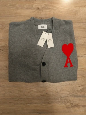 Grå cardigan från Ami med rött hjärta - Grå cardigan från Ami med djup V-ringning och tre knappar framtill. På vänster bröst sitter ett stort rött hjärta med bokstaven A. Tröjan har ribbstickade muddar och nederkant, och är tillverkad i mjukt stickat material.