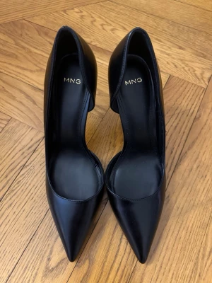 Svarta pumps från MNG med spetsig tå - Oanvända svarta klackskor från MANGO med spetsig tå och hög klack. Skorna har en elegant, öppen sida och är tillverkade i slätt material som ger en klassisk look. Perfekta för dig som gillar minimalistisk och trendig stil. Nypris 550 säljer för 300