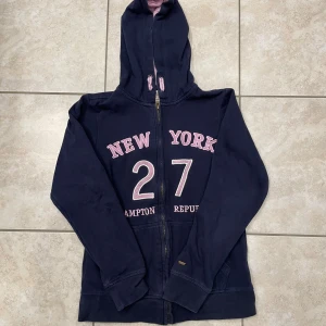 Mörkblå hoodie Hampton Republic - Mörkblå hoodie med dragkedja från Hampton Republic. Tryck i rosa och vitt med texten 'New York 27' på framsidan. Mjuk insida och huva fodrad i ljusrosa tyg. Perfekt för en avslappnad och sportig stil.