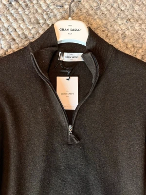 Gran Sasso halfzip - Snygg mörkbrun stickad tröja från Gran Sasso med halv dragkedja och hög krage. Tillverkad i mjuk merinoull som känns lyxig och bekväm. Perfekt för dig som gillar stilrena och klassiska plagg med modern touch.