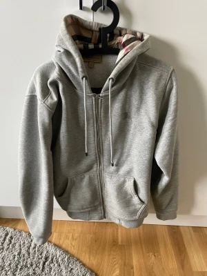 Grå zip hoodie från Burberry - Snygg grå zip hoodie från Burberry med klassiskt rutigt foder i huvan och dragsko. Hoodien har två fickor framtill och är tillverkad i mjukt bomullsmaterial. Perfekt för en avslappnad och stilren look.