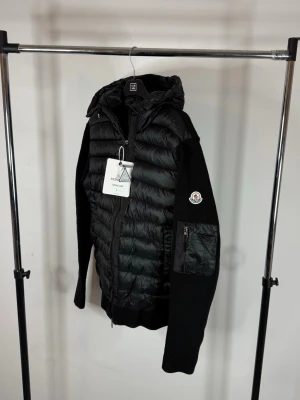 Moncler cardigan - Hybridmodell med quiltad, dunfodrad framsida och huva samt stickade ärmar och rygg – perfekt mix mellan jacka och tröja. Funkar både som vår/höstjacka eller som lager under vinterjackan.  • Märke: Moncler • Färg: Svart • Storlek: L • Skick: Väldigt fint skick (se bilder) • Patch-logo på ärmen • Dragkedja framtill + fickor